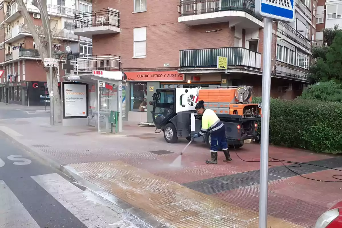 Una persona está limpiando la acera con una hidrolavadora frente a una parada de autobús en una zona urbana.