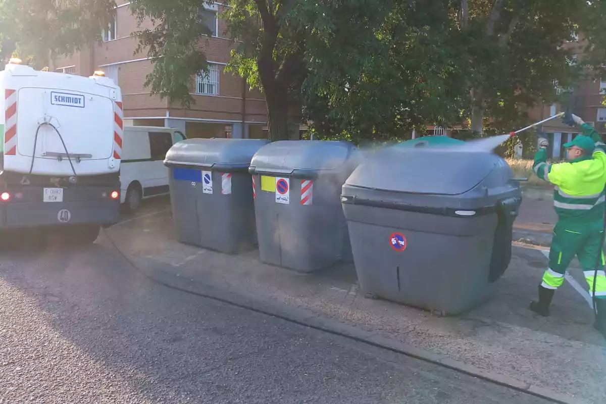 Un trabajador de limpieza con uniforme verde y amarillo está lavando contenedores de basura grises con una manguera de alta presión, mientras un camión de limpieza está estacionado cerca en una calle urbana con árboles y edificios de fondo.