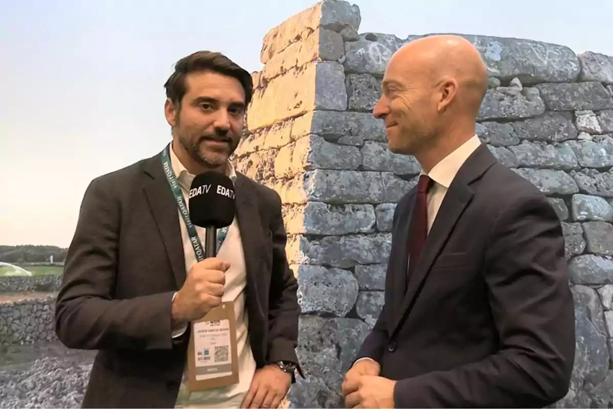 Dos hombres de pie conversando; uno sostiene un micrófono con el logo de EDATV y el otro viste traje y corbata; fondo de un muro de piedra.