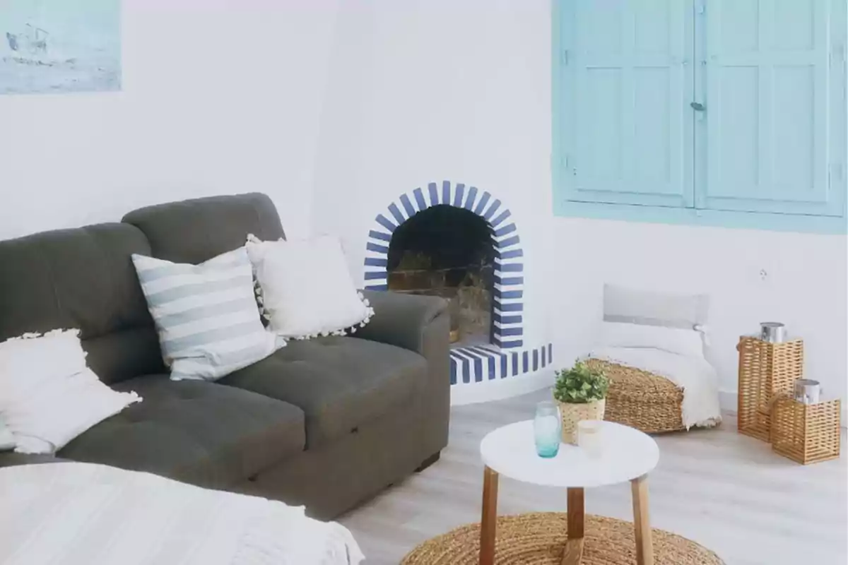 Sala de estar luminosa con sofá gris, cojines blancos, chimenea decorativa, mesa de centro blanca y ventana azul.