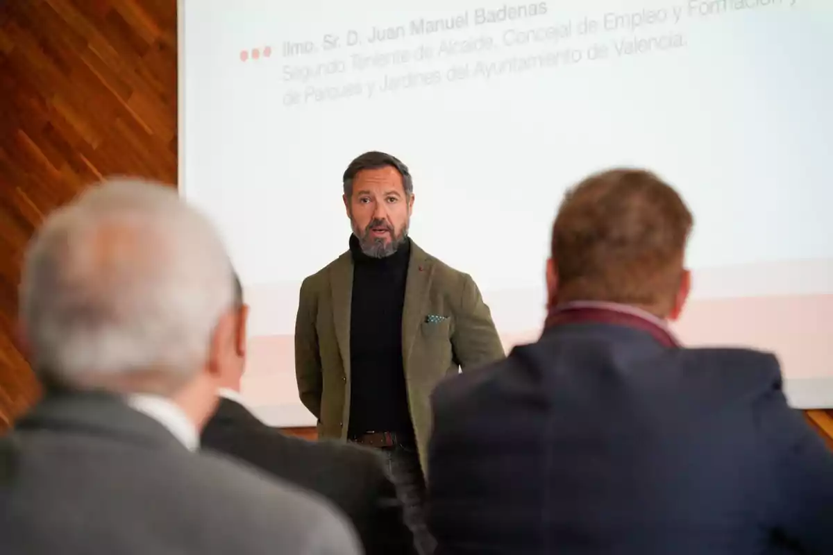 Un hombre de pie frente a una pantalla proyectada, hablando a un grupo de personas en una sala de conferencias.