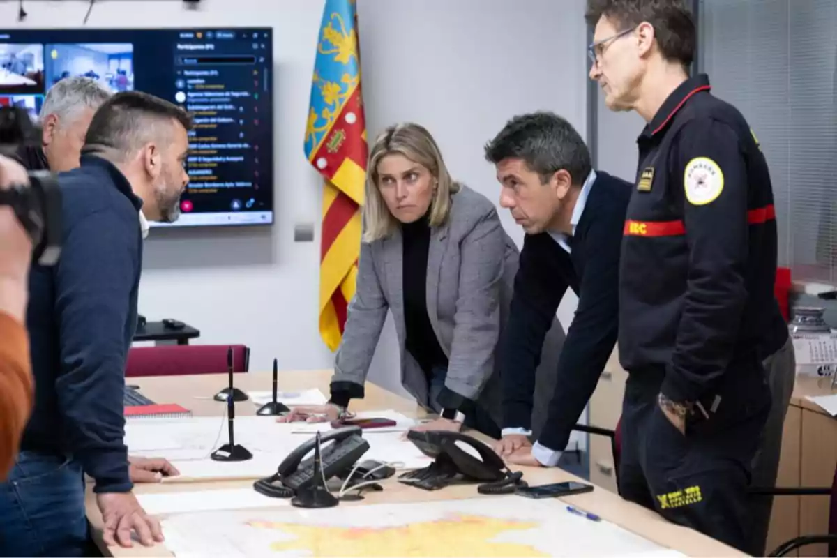 A group of people in a meeting around a table with maps and phones, with a screen and a flag in the background.