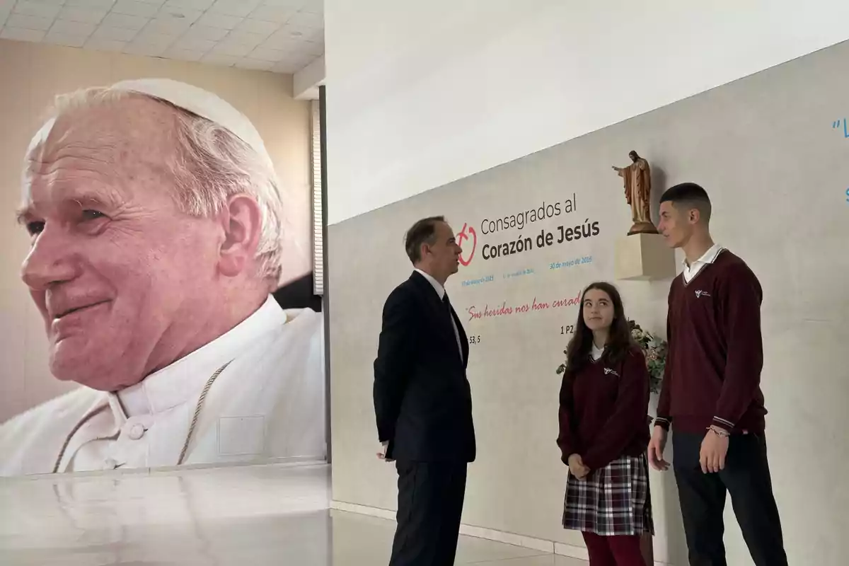 Un hombre y dos estudiantes conversan frente a un mural con la imagen de un papa y una inscripción que dice "Consagrados al Corazón de Jesús".