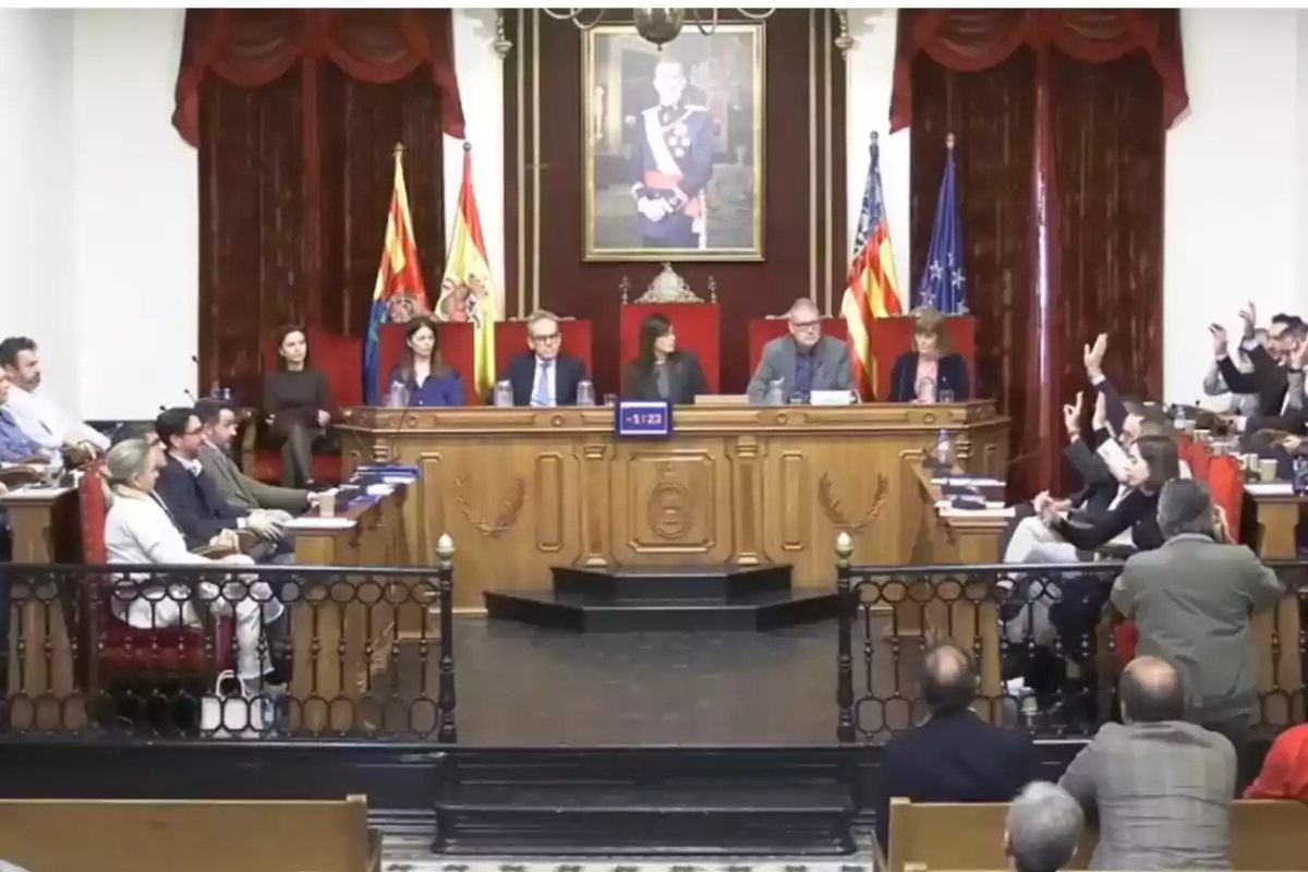 A formal meeting in a government room with several people sitting around a table, some raising their hands, and a portrait on the wall behind the main podium.