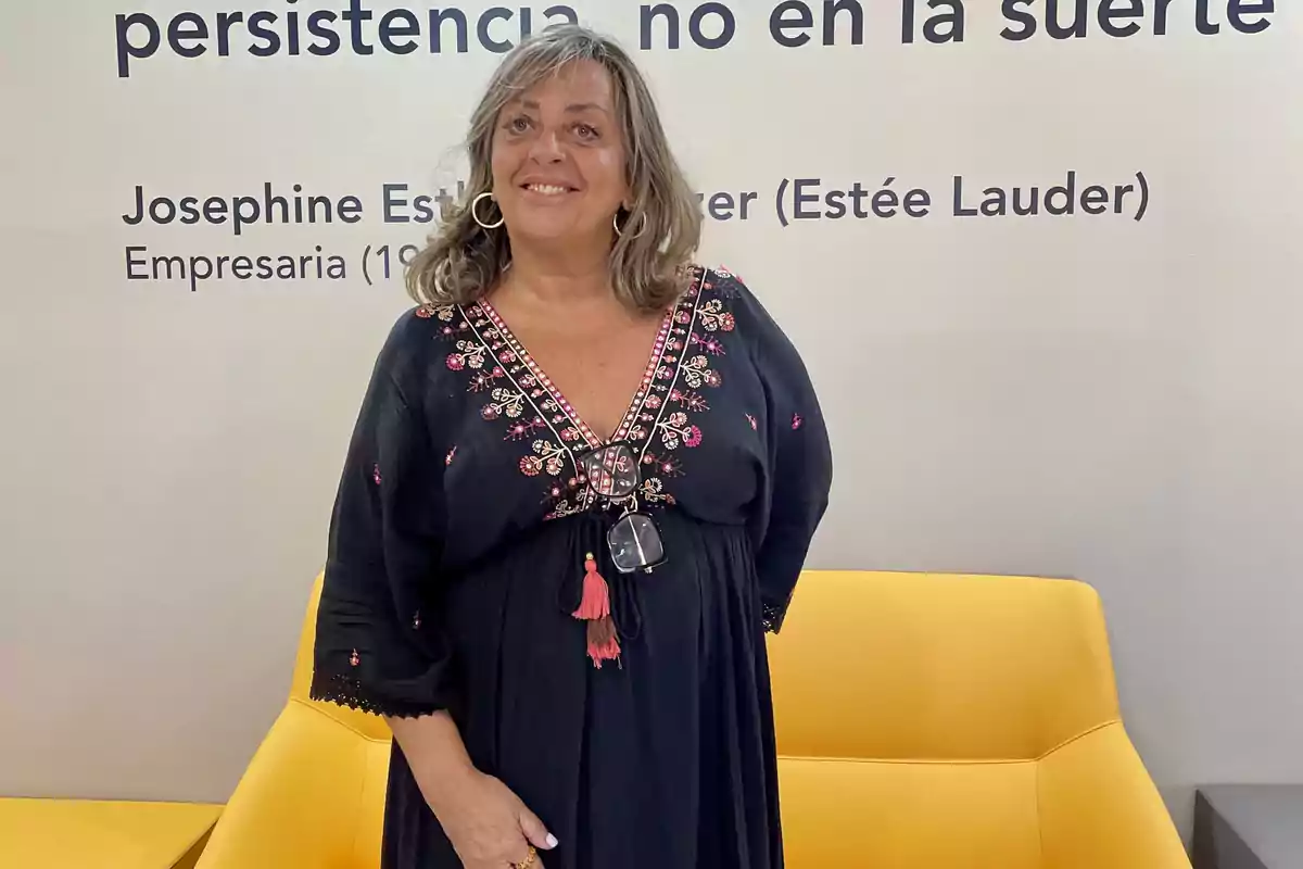 Una mujer sonriente con un vestido negro bordado está de pie frente a una pared con una cita inspiradora de Josephine Esther Mentzer (Estée Lauder).