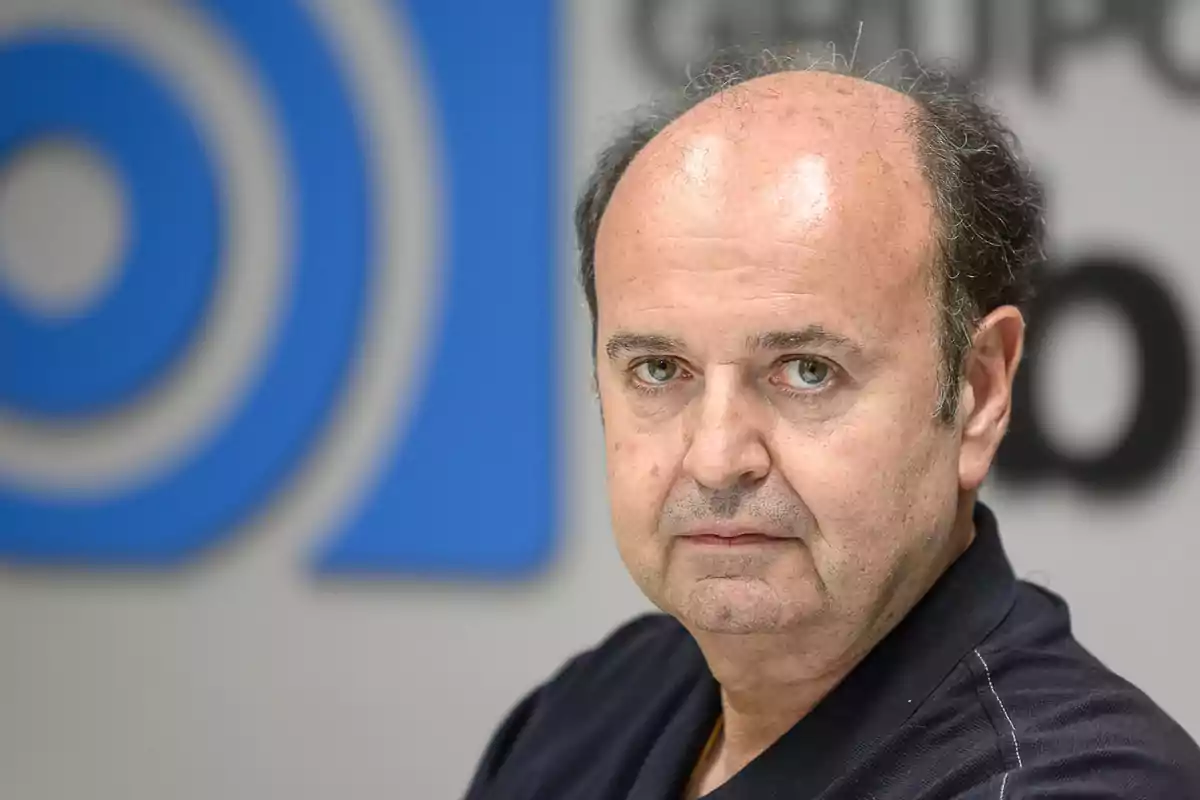 Un hombre de mediana edad con cabello oscuro y escaso mira a la cámara con una expresión seria, con un fondo desenfocado que incluye un logotipo azul.