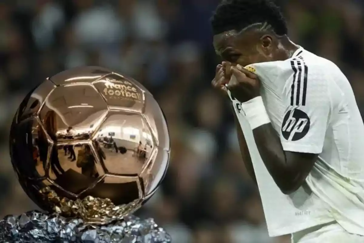 Un jugador de fútbol con camiseta blanca se cubre la boca mientras observa un trofeo dorado en forma de balón.