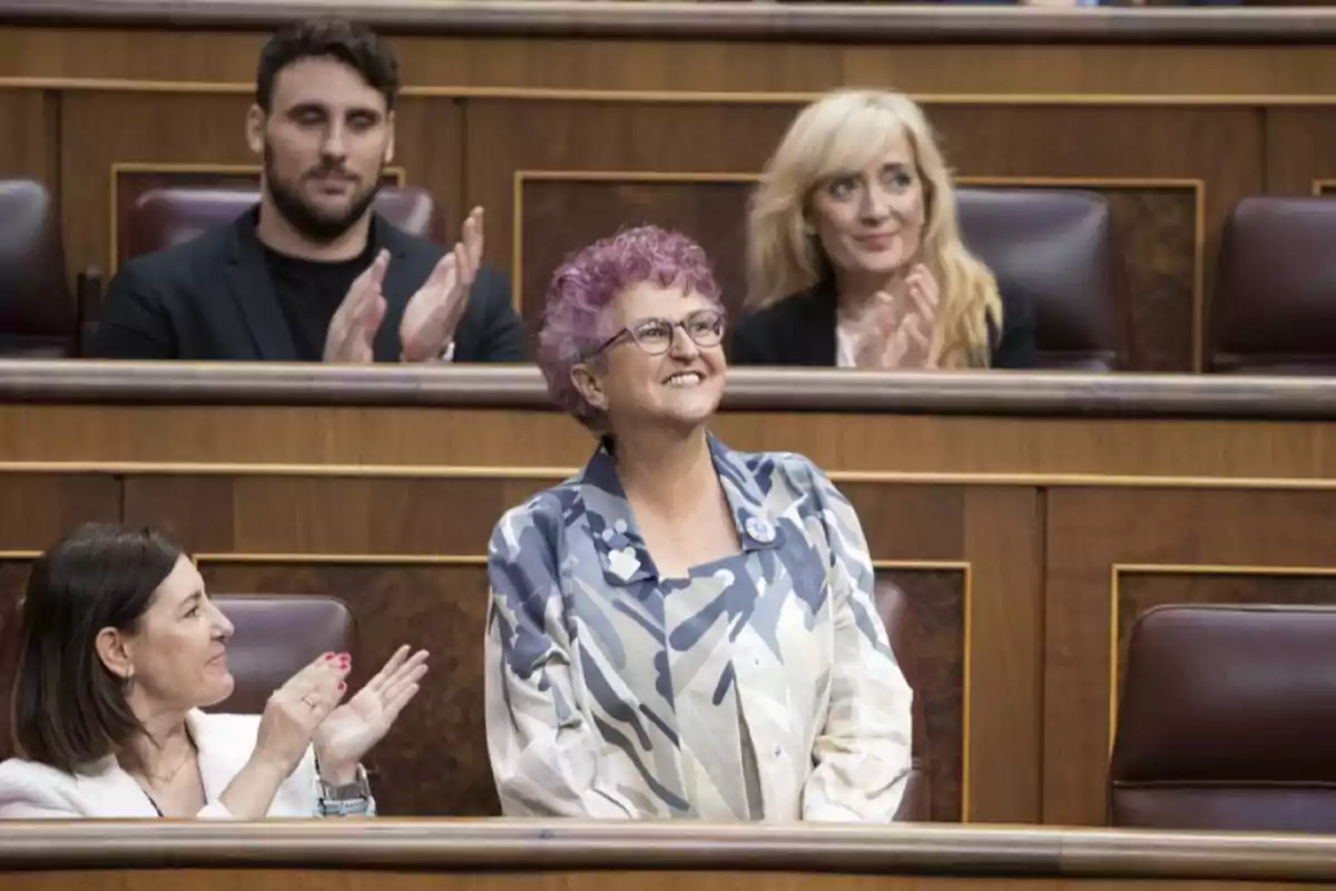 Una mujer de cabello rizado y morado está de pie y sonriendo mientras varias personas a su alrededor la aplauden en un entorno que parece ser un parlamento o una asamblea.