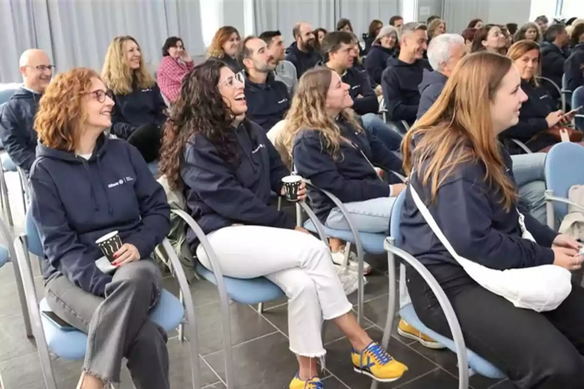 Personas sentadas en una sala de conferencias, vistiendo sudaderas azules y sonriendo mientras escuchan atentamente.