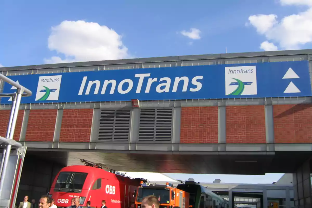 Un cartel de InnoTrans sobre una estructura con trenes y personas debajo, con un cielo azul y nubes en el fondo.