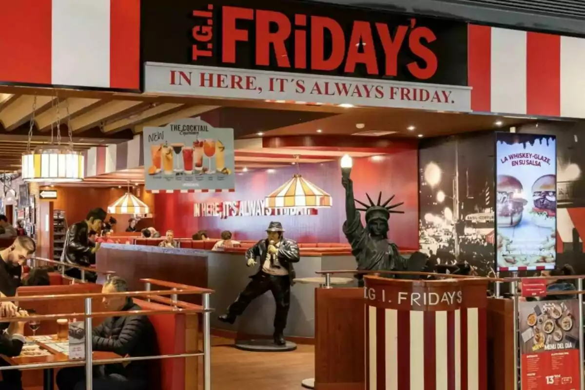 Entrada de un restaurante T.G.I. Friday's con decoración temática, incluyendo una estatua de la Libertad y un maniquí de un músico, donde se observan clientes sentados en mesas disfrutando de su comida. Entrada de un restaurante T.G.I. Friday's con decoración temática, incluyendo una estatua de la Libertad y un maniquí de un músico, donde se observan clientes sentados en mesas disfrutando de su comida.