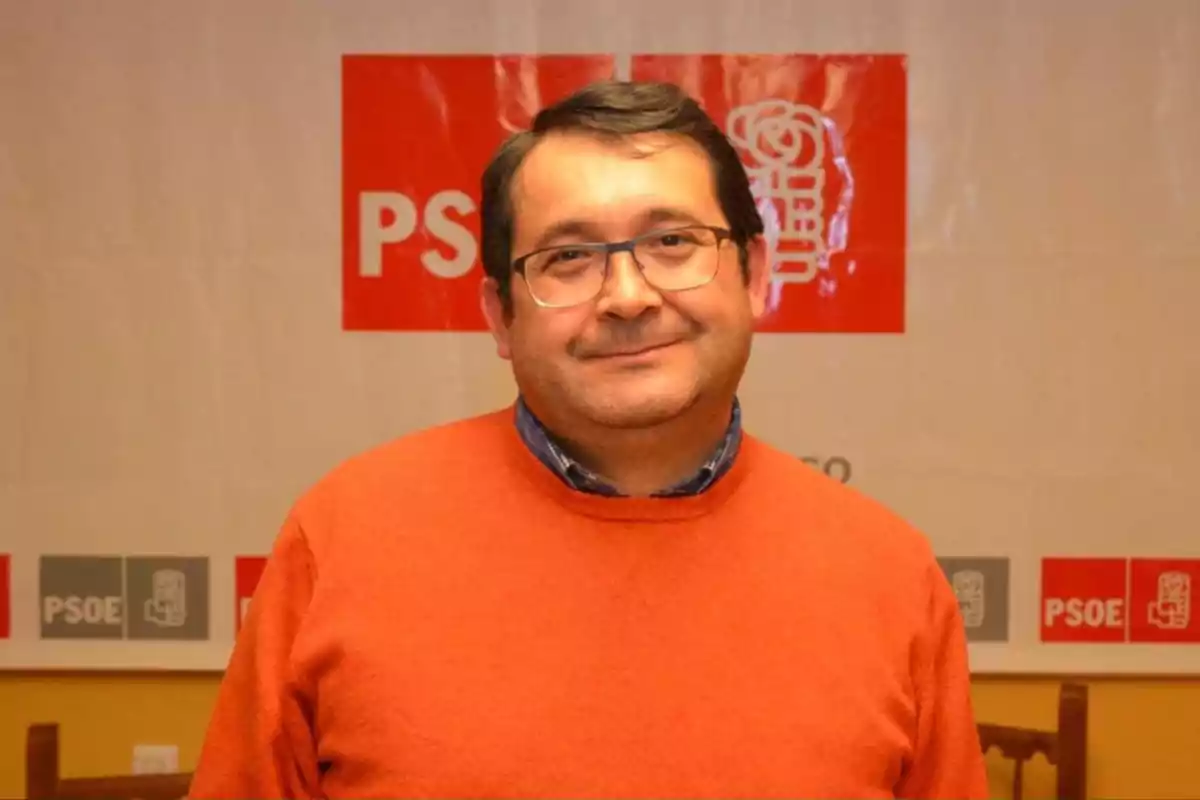 Hombre con gafas y suéter naranja frente a un cartel del PSOE.