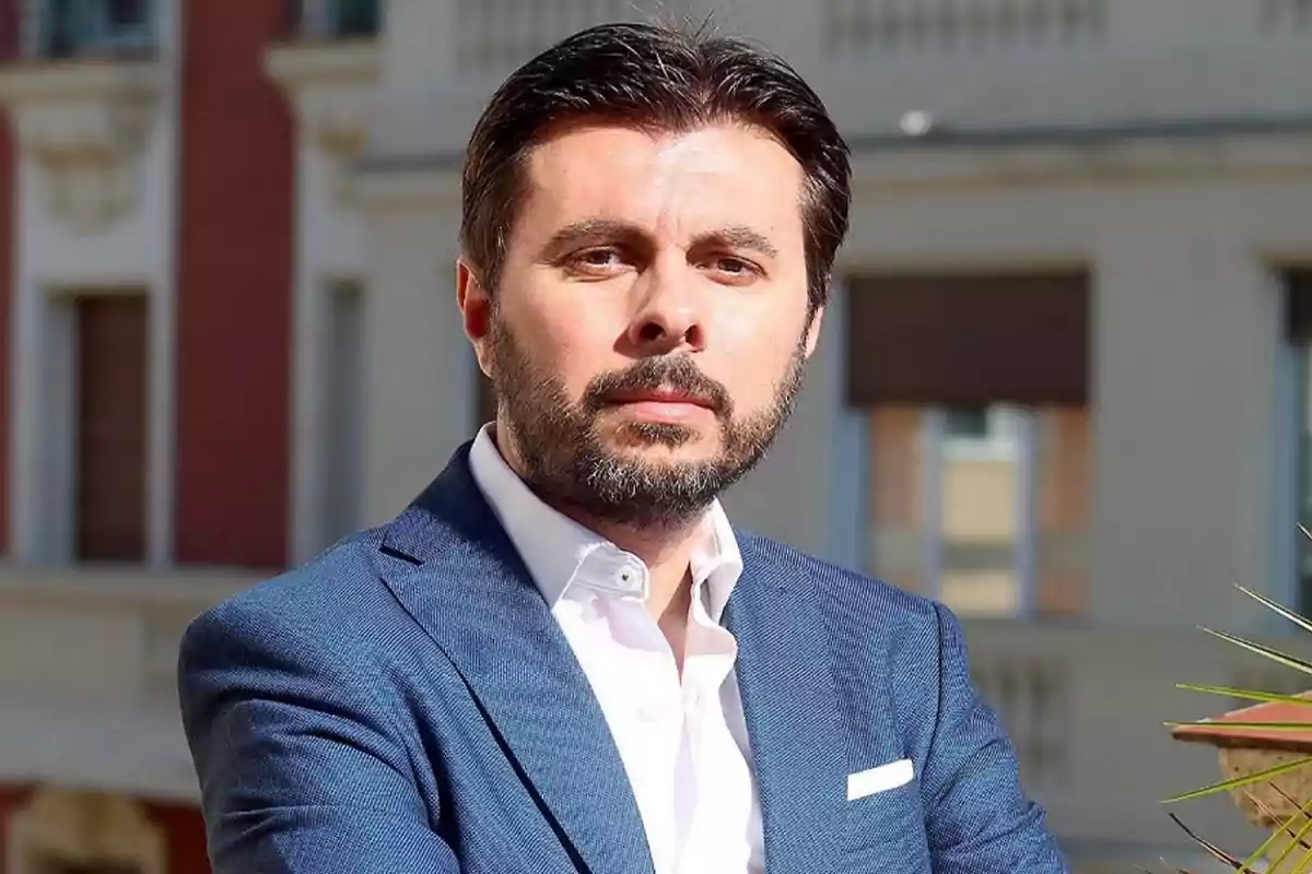 Un hombre con barba y cabello oscuro lleva un traje azul y camisa blanca, posando al aire libre frente a un edificio.