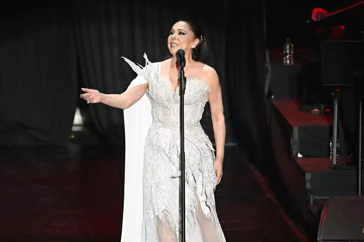 Isabel Pantoja con un vestido blanco elegante canta en un escenario iluminado.