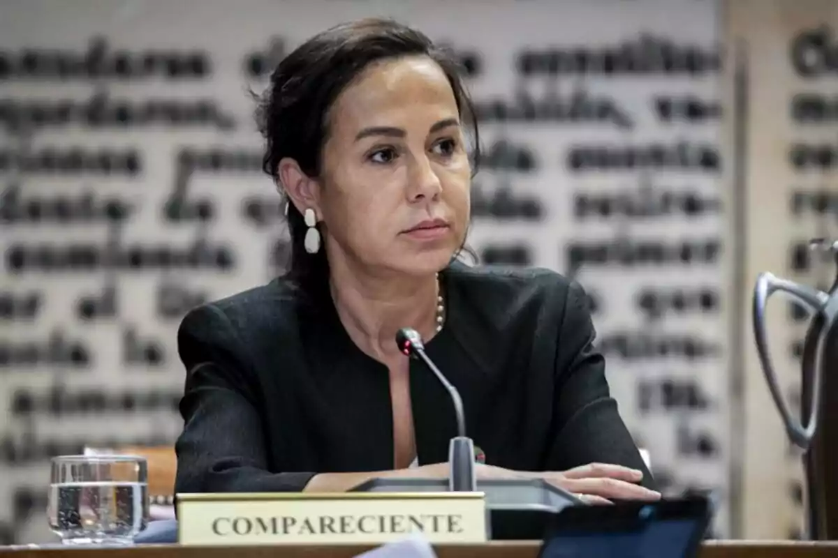 Una mujer sentada en una mesa con un micrófono frente a ella y un cartel que dice Una mujer sentada en una mesa con un micrófono frente a ella y un cartel que dice