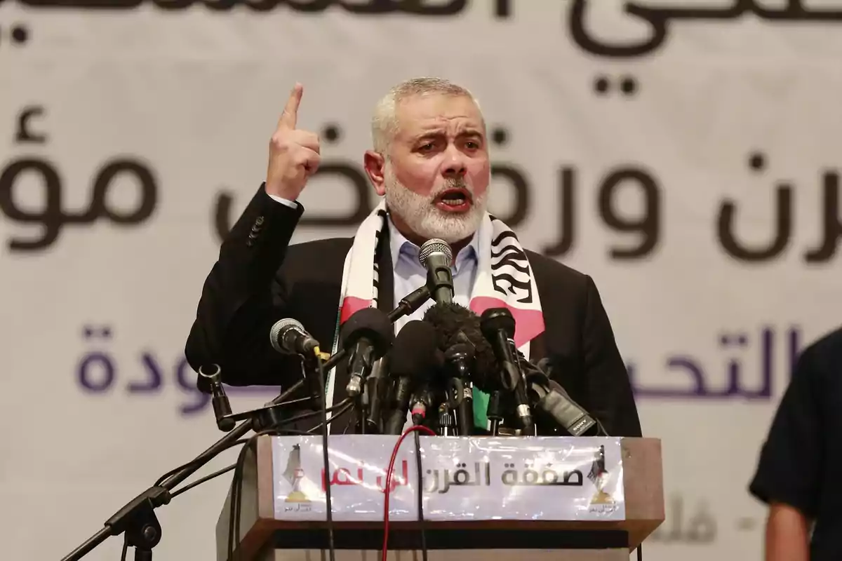 Un hombre con barba blanca y cabello corto, vestido con traje oscuro y una bufanda con los colores de la bandera palestina, habla en un podio con varios micrófonos frente a él, levantando un dedo mientras se dirige al público.