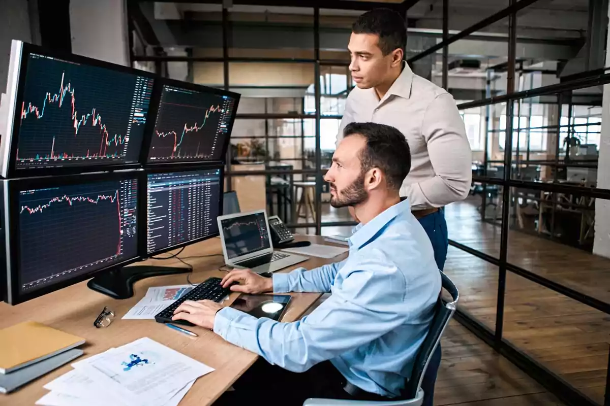 Dos hombres trabajando en una oficina con múltiples pantallas de computadora mostrando gráficos financieros.