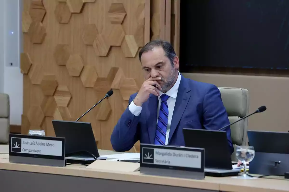 Un hombre en traje azul está sentado en una mesa de conferencias, pensativo, con una mano en el rostro y rodeado de computadoras portátiles y micrófonos.