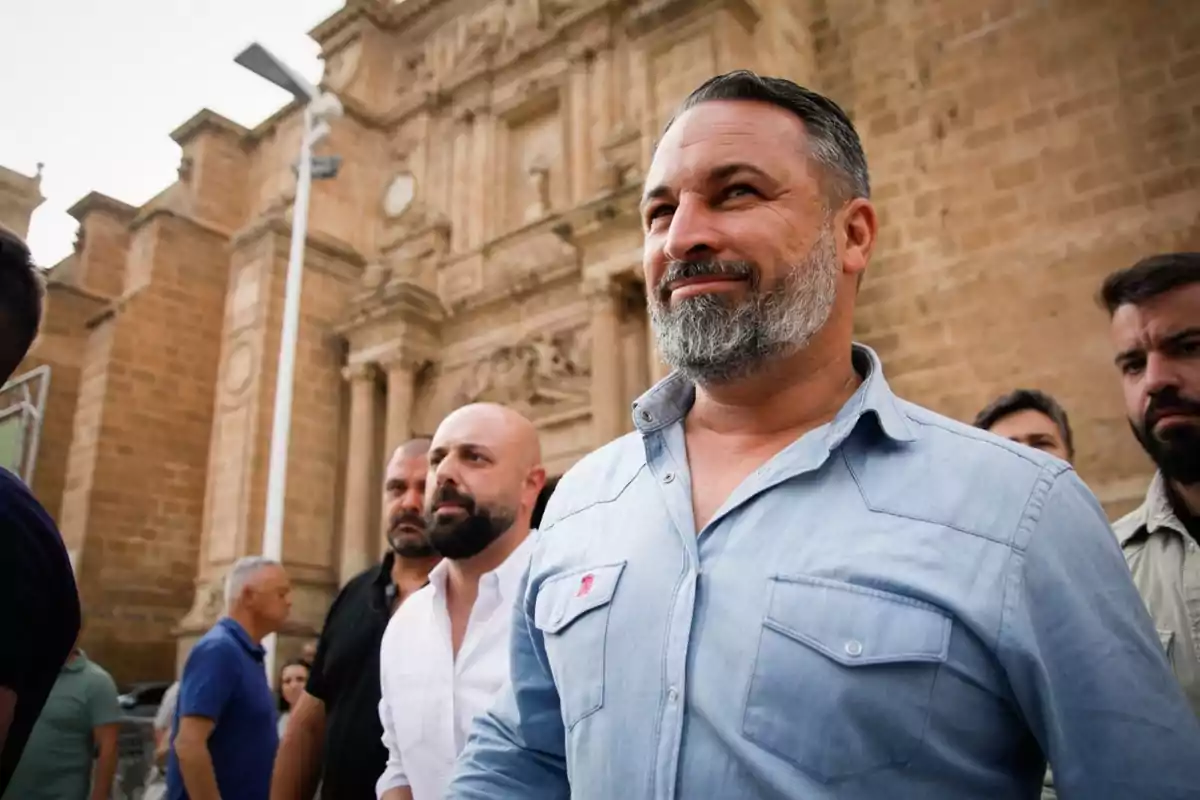 Hombre con barba gris y camisa azul claro sonriendo frente a un edificio histórico de piedra, acompañado de varias personas.