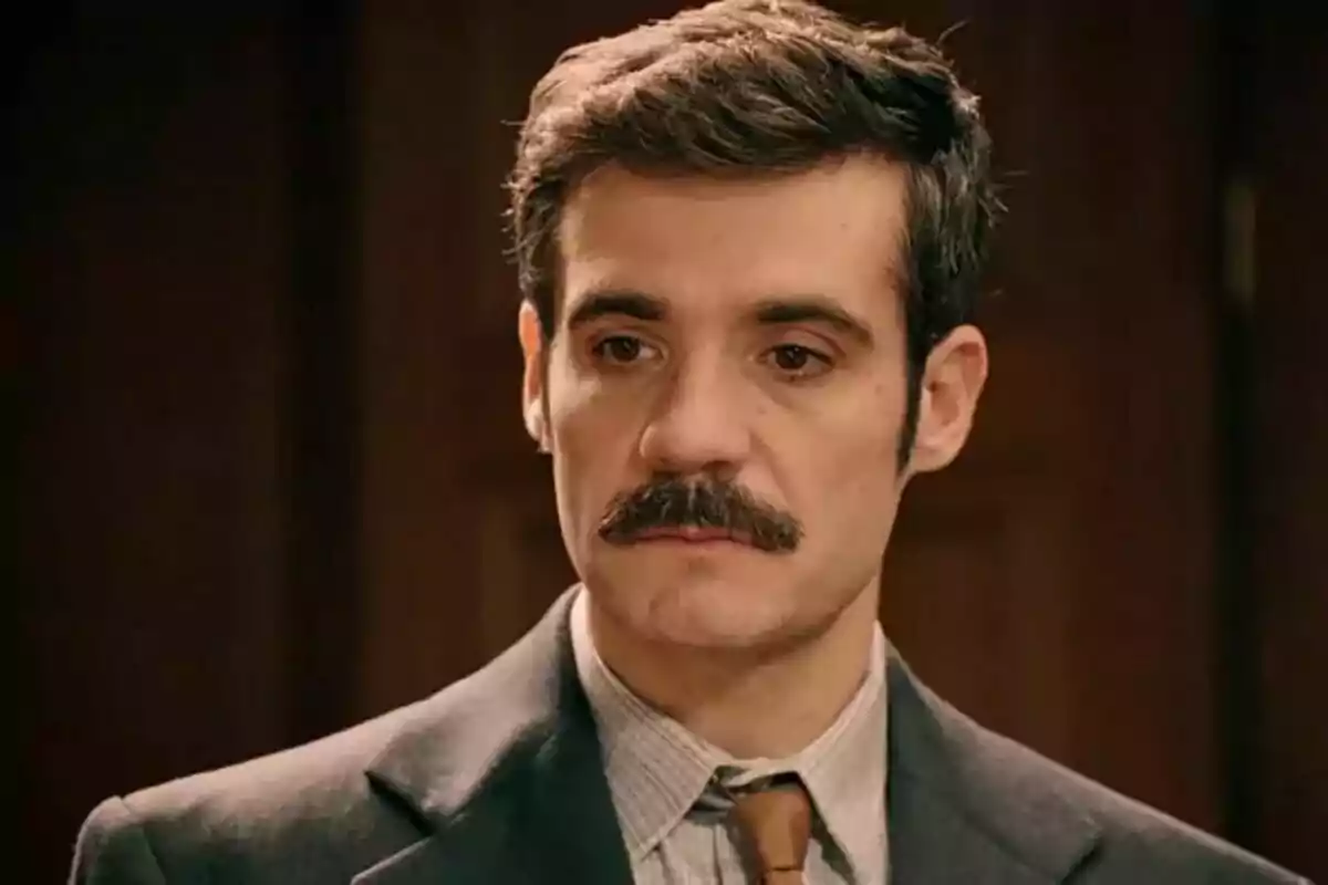 Plano corto del actor Javier Beltrán con bigote y traje mira pensativo hacia adelante.