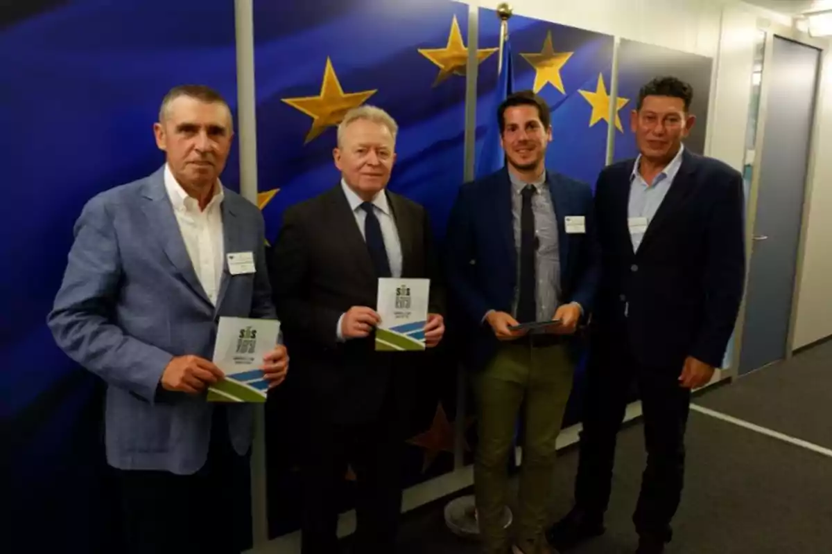 Cuatro hombres posan para una foto frente a una bandera de la Unión Europea. Tres de ellos sostienen folletos en sus manos. Están vestidos con trajes formales y parecen estar en un entorno de oficina o conferencia.