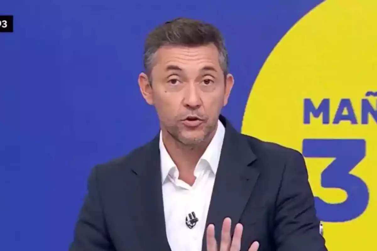 Un hombre con traje oscuro y camisa blanca está hablando frente a un fondo azul y amarillo con texto. Un hombre con traje oscuro y camisa blanca está hablando frente a un fondo azul y amarillo con texto.