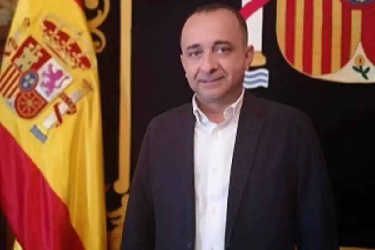 Un hombre de pie con traje oscuro y camisa blanca frente a una bandera de España. Un hombre de pie con traje oscuro y camisa blanca frente a una bandera de España.