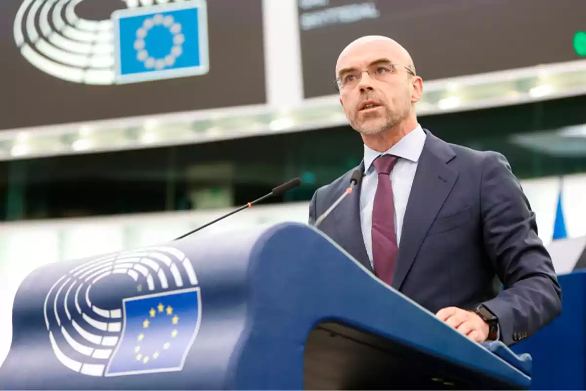 Hombre de traje hablando en un podio con el logo de la Unión Europea en un entorno institucional.