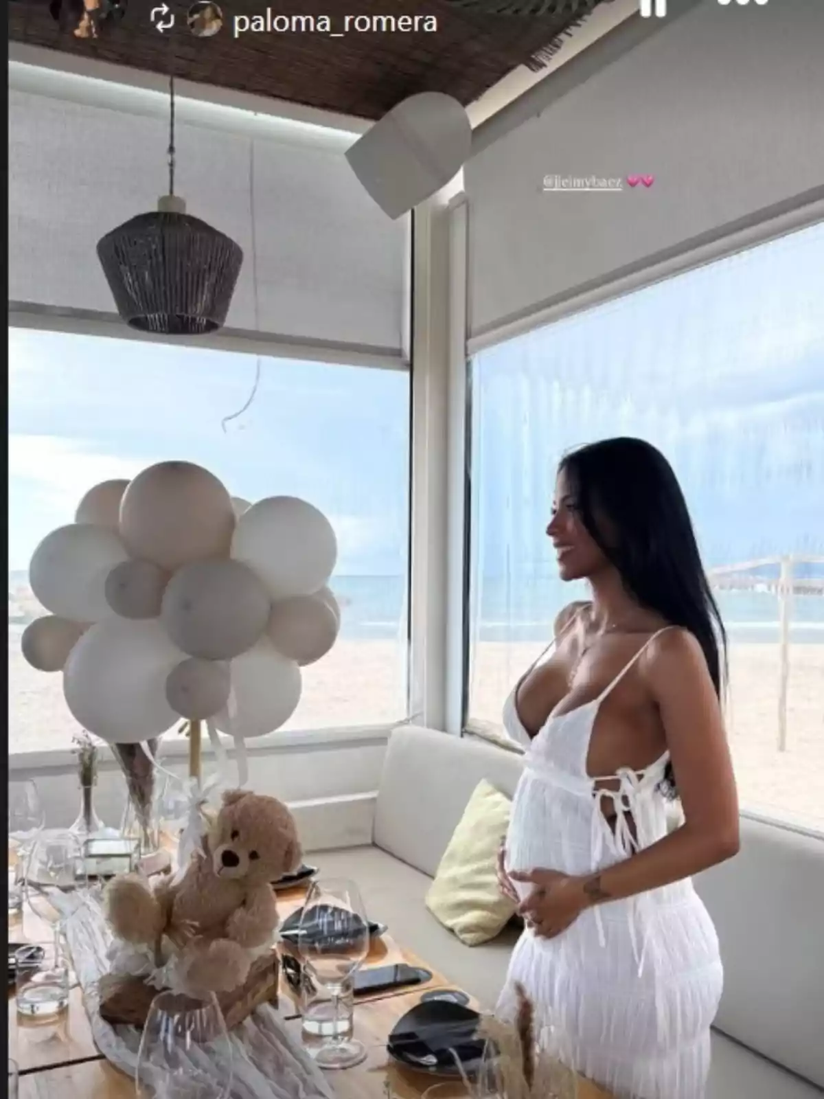 Jeimy Báez con un vestido blanco está de pie junto a una mesa decorada con globos blancos presume de barriga de embarazada Jeimy Báez con un vestido blanco está de pie junto a una mesa decorada con globos blancos presume de barriga de embarazada