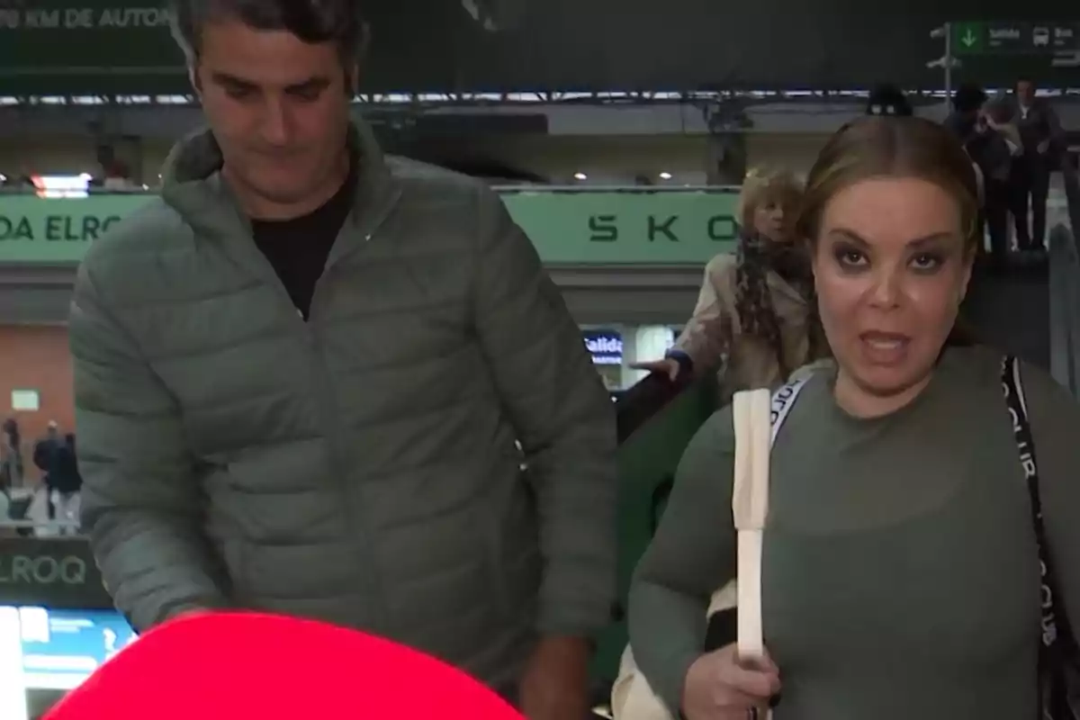 Jesulín de Ubrique y María José Campanario en una estación de tren, una mujer con una camiseta verde y una bolsa blanca y un hombre con una chaqueta verde, con gente y carteles en el fondo. Jesulín de Ubrique y María José Campanario en una estación de tren, una mujer con una camiseta verde y una bolsa blanca y un hombre con una chaqueta verde, con gente y carteles en el fondo.