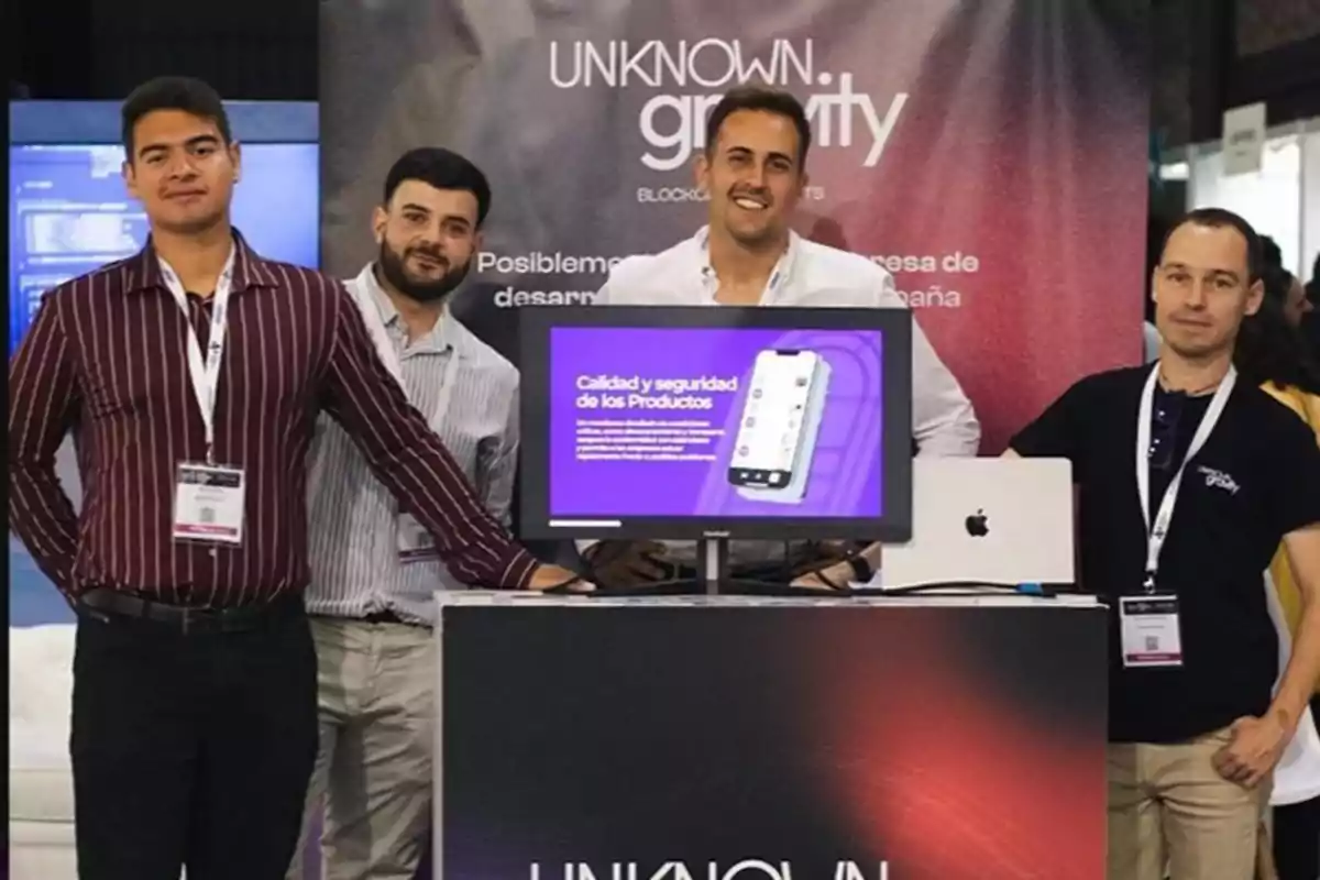 Cuatro hombres posan frente a un stand de exhibición con un monitor que muestra información sobre la calidad y seguridad de los productos, con un fondo que dice "Unknown Gravity".