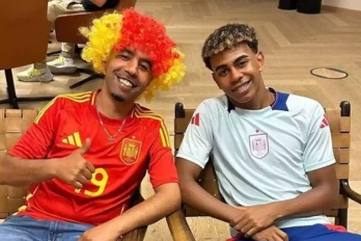 Dos personas sentadas, una con una peluca de colores rojo y amarillo y una camiseta de la selección española de fútbol, y la otra con una camiseta de entrenamiento de la misma selección.