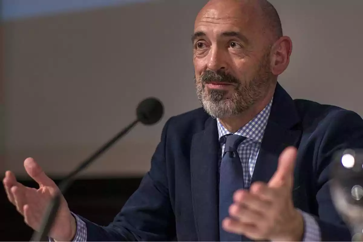 Hombre calvo con barba hablando en un micrófono durante una conferencia, vestido con traje azul y camisa a cuadros.