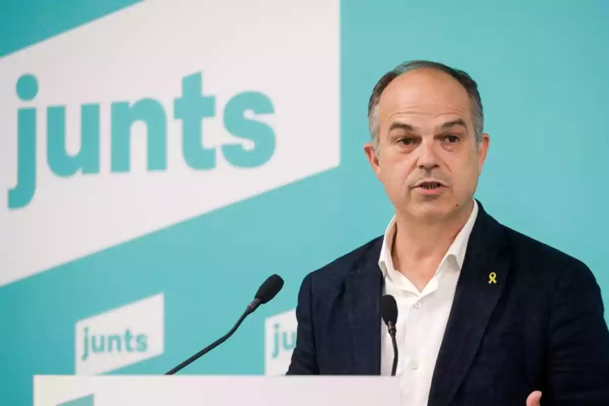 Hombre hablando en una conferencia de prensa con un fondo azul que tiene el logo de "junts".