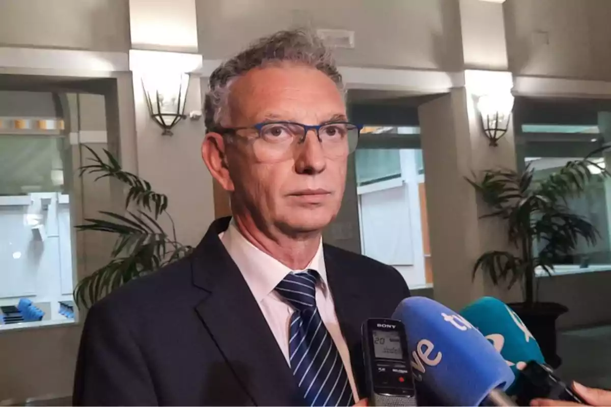 Un hombre con gafas y traje oscuro es entrevistado por varios medios de comunicación en un entorno interior iluminado. Un hombre con gafas y traje oscuro es entrevistado por varios medios de comunicación en un entorno interior iluminado.