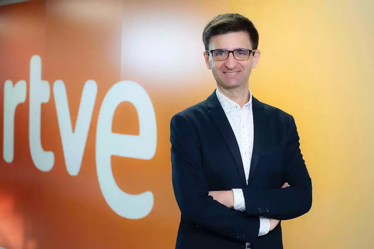 Un hombre con gafas y traje oscuro sonríe frente a un fondo naranja con el logotipo de RTVE.