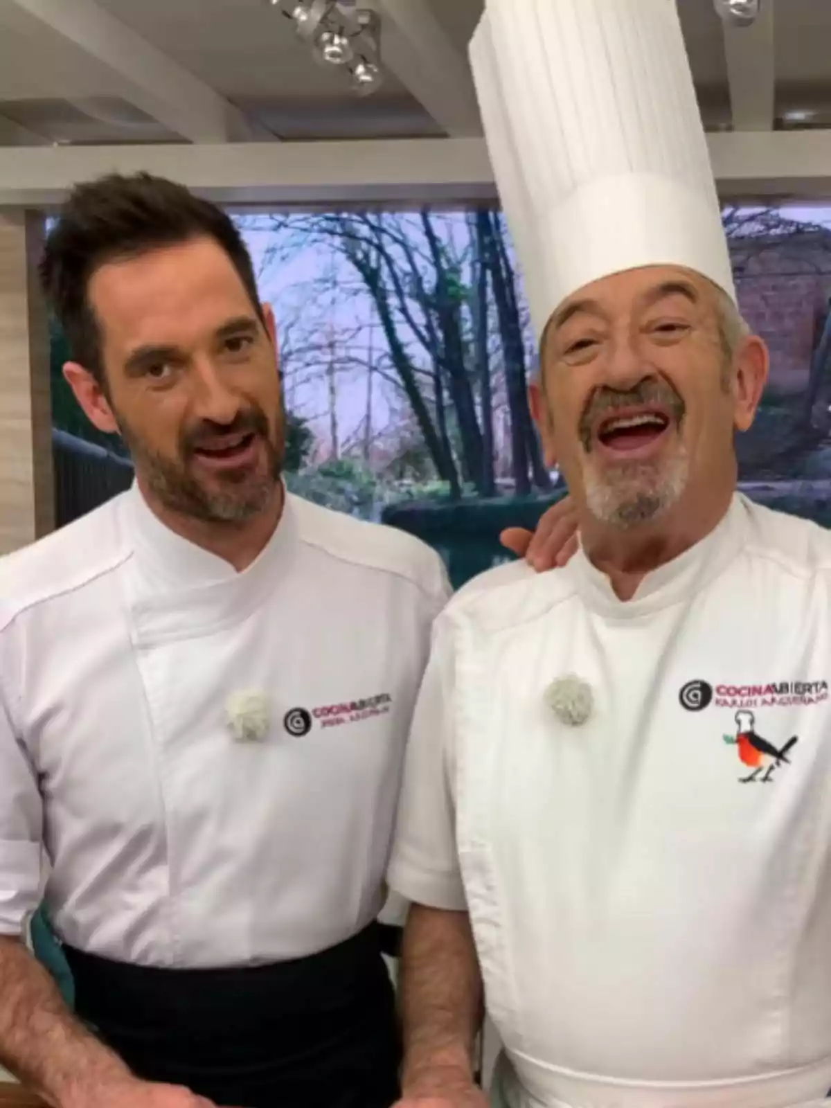 Dos chefs -que son Joseba Arguiñano y Karlos Arguiñano- sonríen juntos en una cocina, ambos llevan uniforme blanco y uno de ellos lleva un gorro alto de chef Dos chefs -que son Joseba Arguiñano y Karlos Arguiñano- sonríen juntos en una cocina, ambos llevan uniforme blanco y uno de ellos lleva un gorro alto de chef