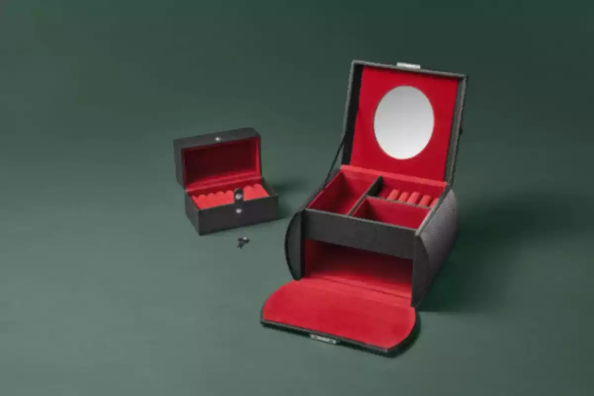 Caja de joyería negra con interior rojo y espejo en la tapa abierta junto a una caja más pequeña sobre una superficie verde. Caja de joyería negra con interior rojo y espejo en la tapa abierta junto a una caja más pequeña sobre una superficie verde.