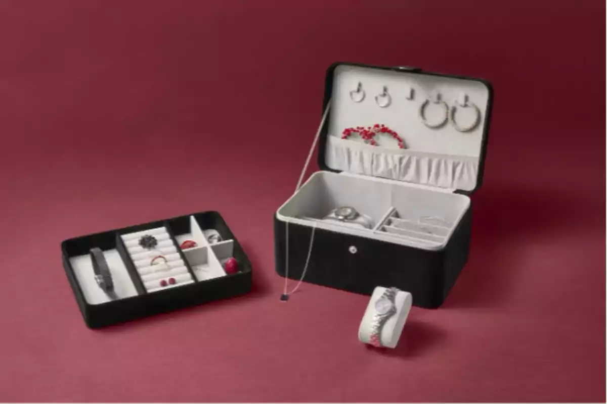 Estuche de joyería abierto mostrando varios compartimentos con anillos, pulseras y aretes sobre un fondo rojo. Estuche de joyería abierto mostrando varios compartimentos con anillos, pulseras y aretes sobre un fondo rojo.
