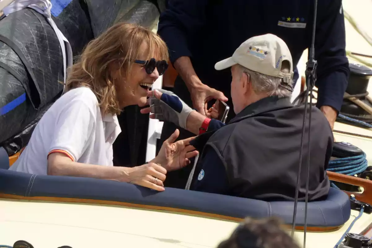 A smiling woman with sunglasses interacts with Juan Carlos I on a boat, surrounded by nautical equipment. A smiling woman with sunglasses interacts with Juan Carlos I on a boat, surrounded by nautical equipment.