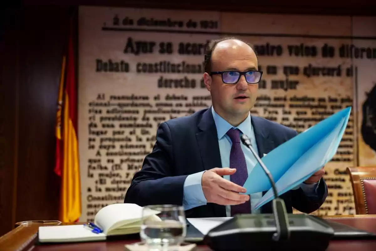 Un hombre con gafas y traje sostiene una carpeta azul en una sala de conferencias con un fondo de texto histórico y una bandera de España. Un hombre con gafas y traje sostiene una carpeta azul en una sala de conferencias con un fondo de texto histórico y una bandera de España.