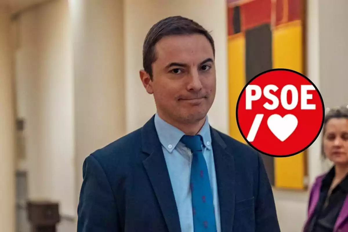 Un hombre con traje y corbata azul está de pie en un pasillo con un logotipo rojo del PSOE a su lado.