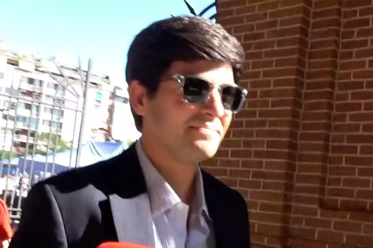 Juan Urquijo con gafas de sol y saco oscuro caminando al aire libre junto a una pared de ladrillos Juan Urquijo con gafas de sol y saco oscuro caminando al aire libre junto a una pared de ladrillos