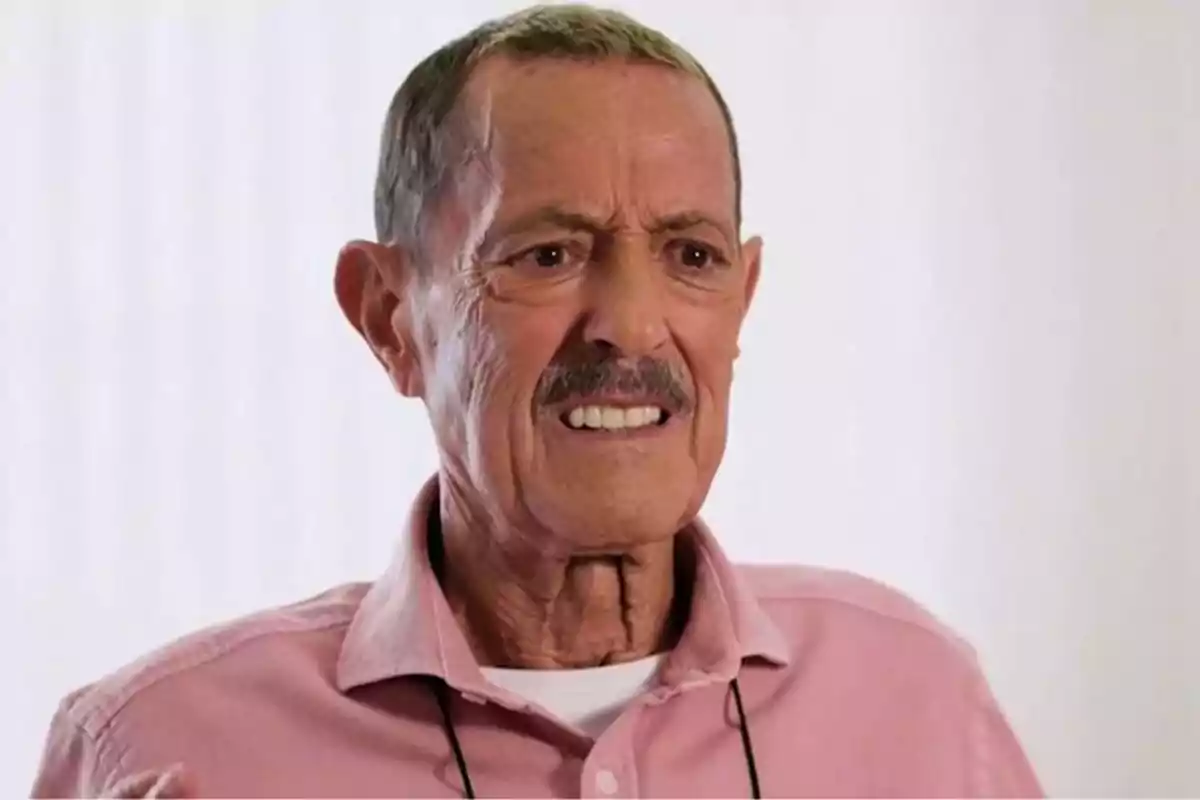 Un hombre mayor con bigote y cabello corto, vistiendo una camisa rosa, con una expresión facial seria.