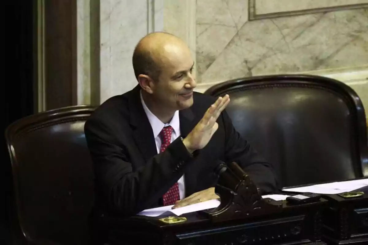 Un hombre calvo con traje y corbata roja, sentado en un escritorio de madera oscura, levantando la mano y sonriendo.