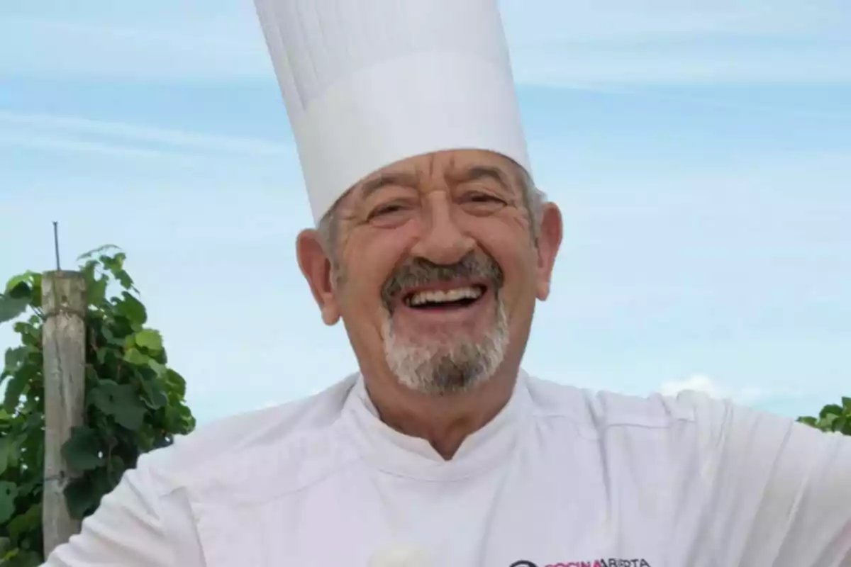 Karlos Arguiñano sonriente con un gorro de cocinero blanco y una chaqueta de chef blanca, con un fondo de cielo azul y vegetación en Cocina abierta de Karlos Arguiñano.