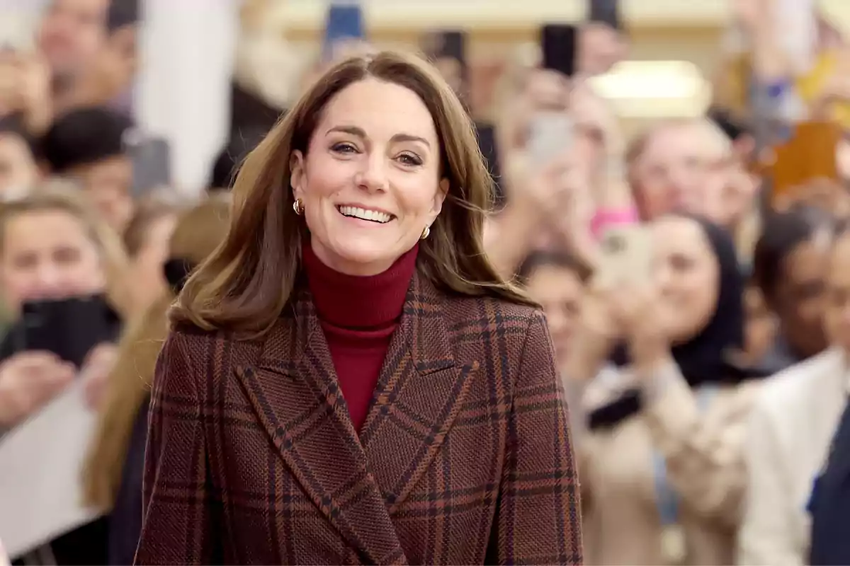 Kate Middleton smiling in a brown plaid coat and a dark red sweater, surrounded by people holding mobile phones. Kate Middleton smiling in a brown plaid coat and a dark red sweater, surrounded by people holding mobile phones.