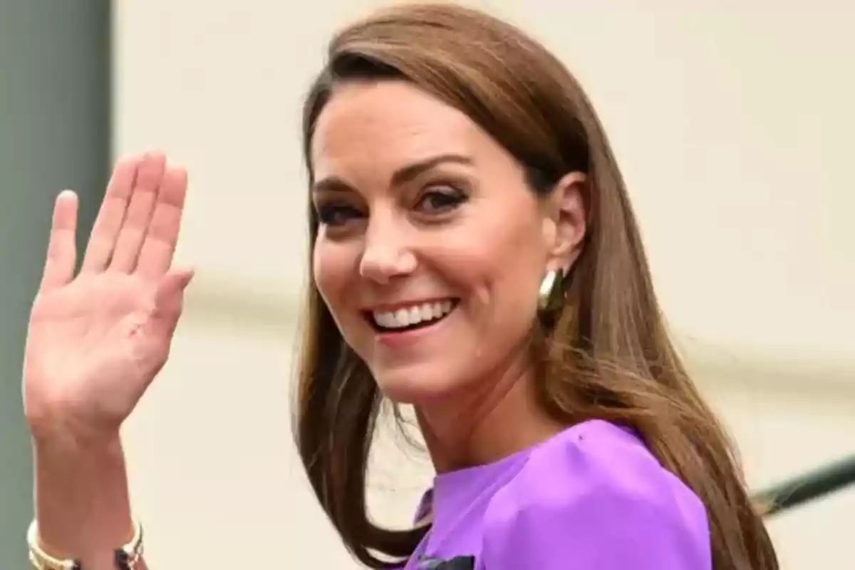 Persona sonriendo y saludando con la mano levantada, lleva una blusa morada y aretes dorados.