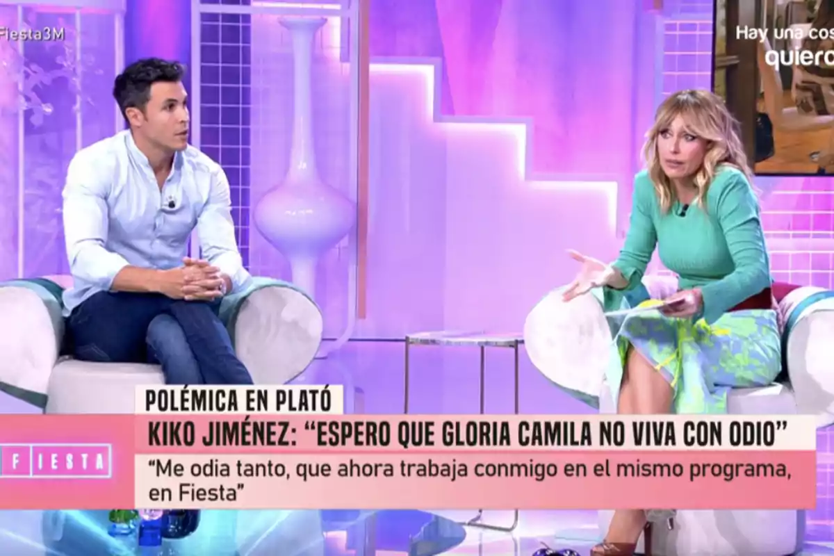 En un plató de televisión, Kiko Jiménez y Emma García están sentadas en sillones blancos, conversando en un ambiente iluminado con luces rosadas y moradas, mientras en la parte inferior de la imagen se muestra un texto sobre una polémica relacionada con un programa llamado En un plató de televisión, Kiko Jiménez y Emma García están sentadas en sillones blancos, conversando en un ambiente iluminado con luces rosadas y moradas, mientras en la parte inferior de la imagen se muestra un texto sobre una polémica relacionada con un programa llamado