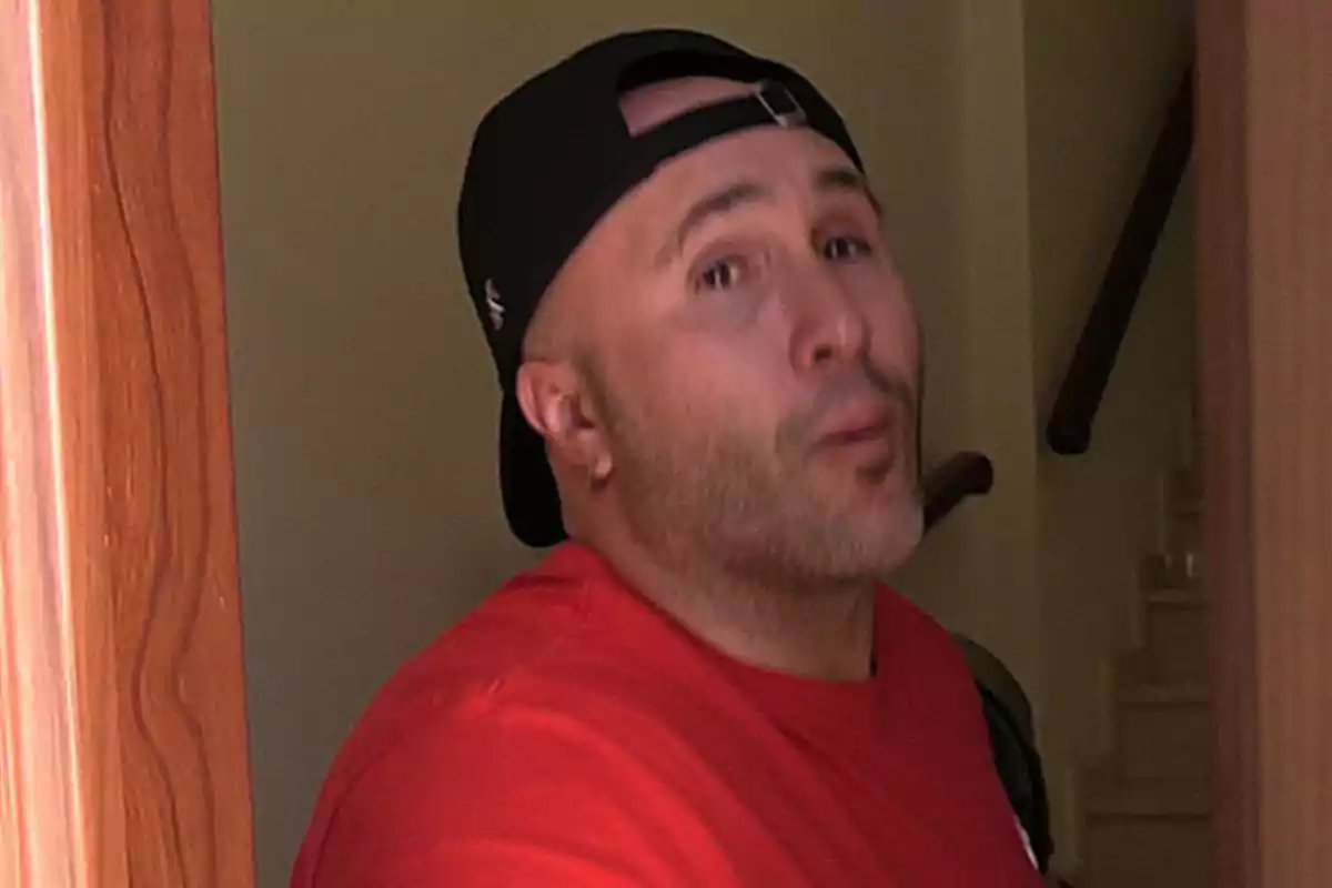 Kiko Rivera con gorra negra al revés y camiseta roja está de pie en el umbral de una puerta de madera. Kiko Rivera con gorra negra al revés y camiseta roja está de pie en el umbral de una puerta de madera.